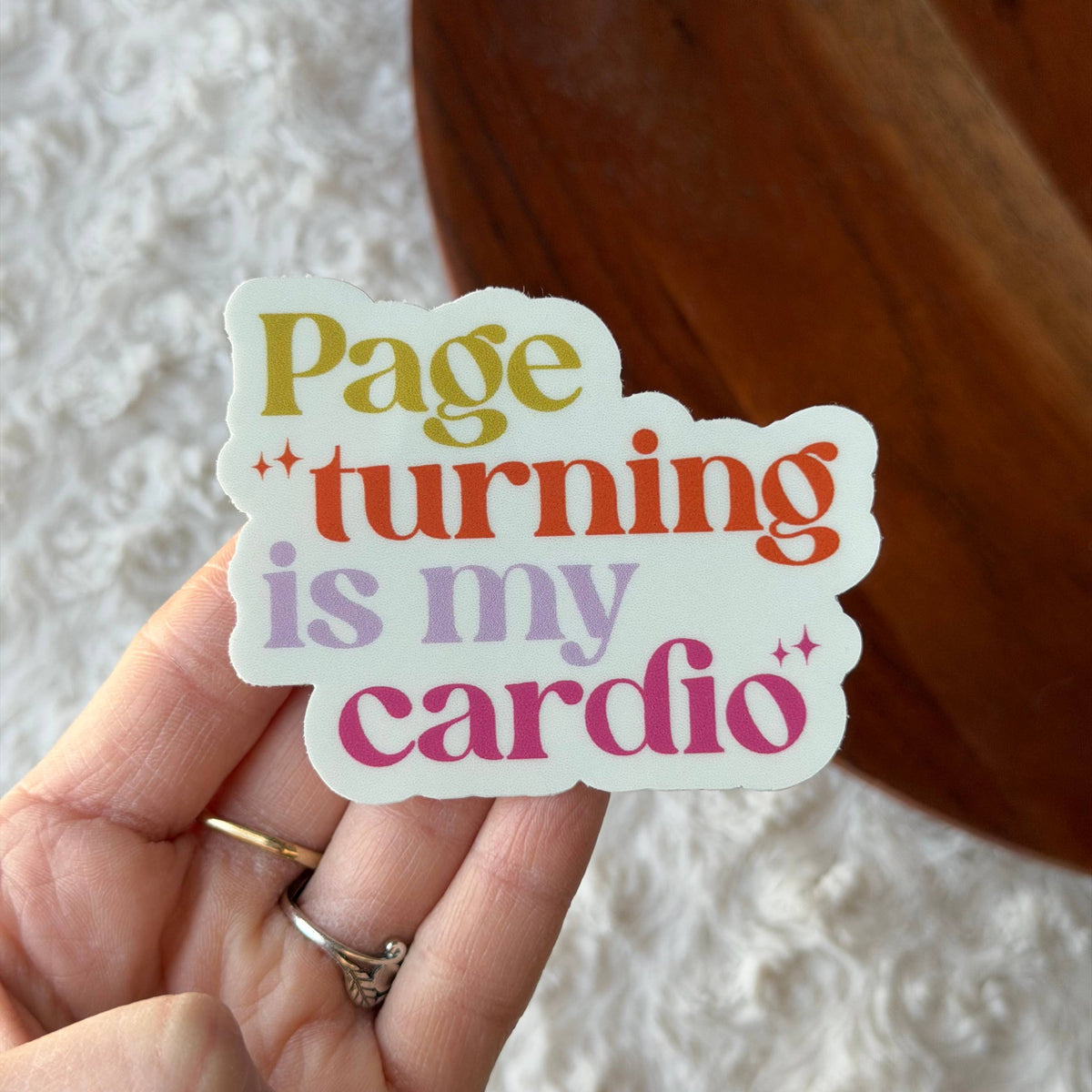 Hand holding a colorful sticker with text 'Page turning is my cardio' against a textured white background.