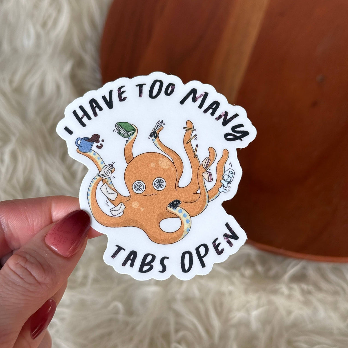 A hand with red painted nails holds a die-cut sticker featuring an orange cartoon octopus with multiple colored browser tabs and the text 'I HAVE TOO MANY TABS OPEN'. The background shows a soft white faux fur surface and a warm brown wooden object.