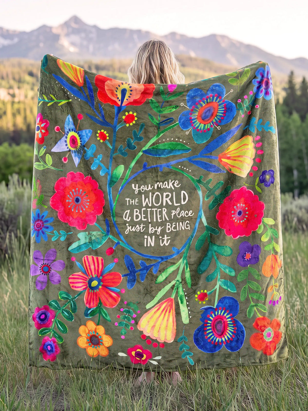 WOMAN HOLDING MULTI COLOR BLANKET WITH FLOWERS AND THE WORDS YOU MAKE THE WORLD A BETTER PLACE JUST BY BEING IN IT