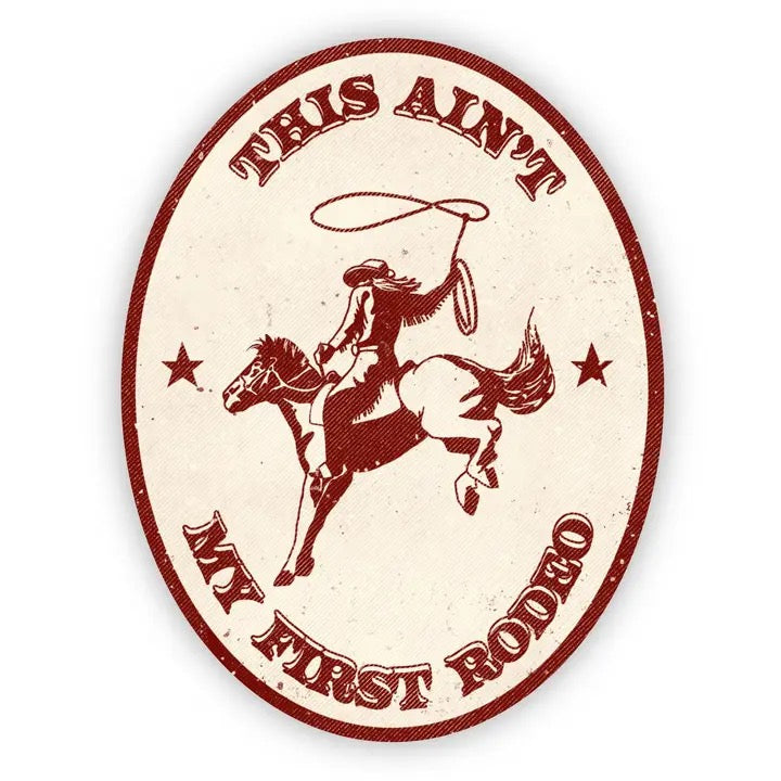 An oval-shaped sticker featuring a cowboy riding a bucking horse while throwing a lasso. The text reads 'THIS AIN'T MY FIRST RODEO' in a western-style font, with two stars on either side of the rider.