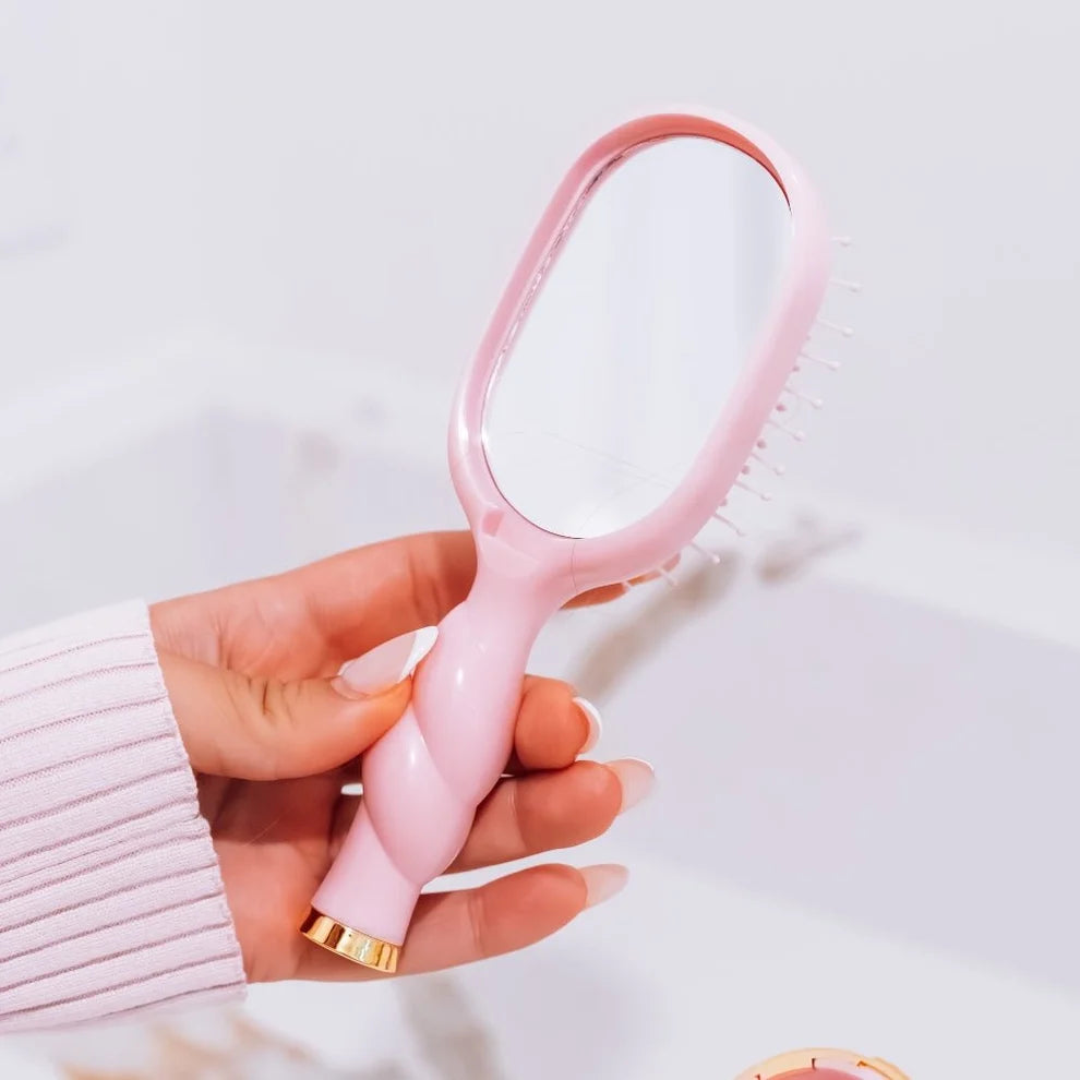 A hand holding a pink oval hairbrush with a mirror on the back and soft white bristles, against a bright white background.