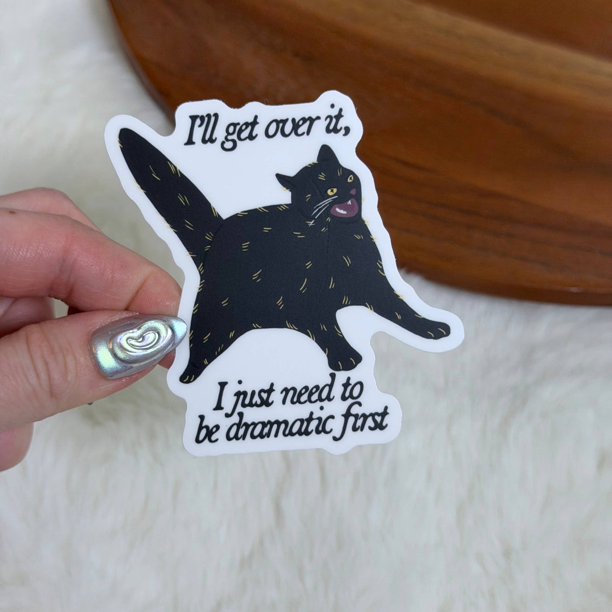 Sticker with a black cat illustration and humorous text held by a hand.