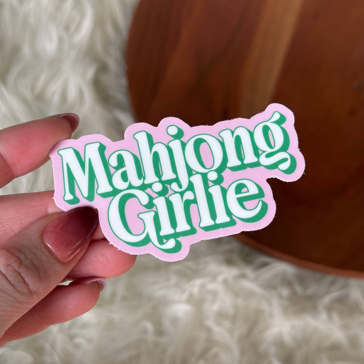 Sticker with 'Mahjong Girlie' text held by a hand against a soft background