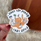 A hand with red painted nails holds a die-cut sticker featuring an orange cartoon octopus with multiple colored browser tabs and the text 'I HAVE TOO MANY TABS OPEN'. The background shows a soft white faux fur surface and a warm brown wooden object.