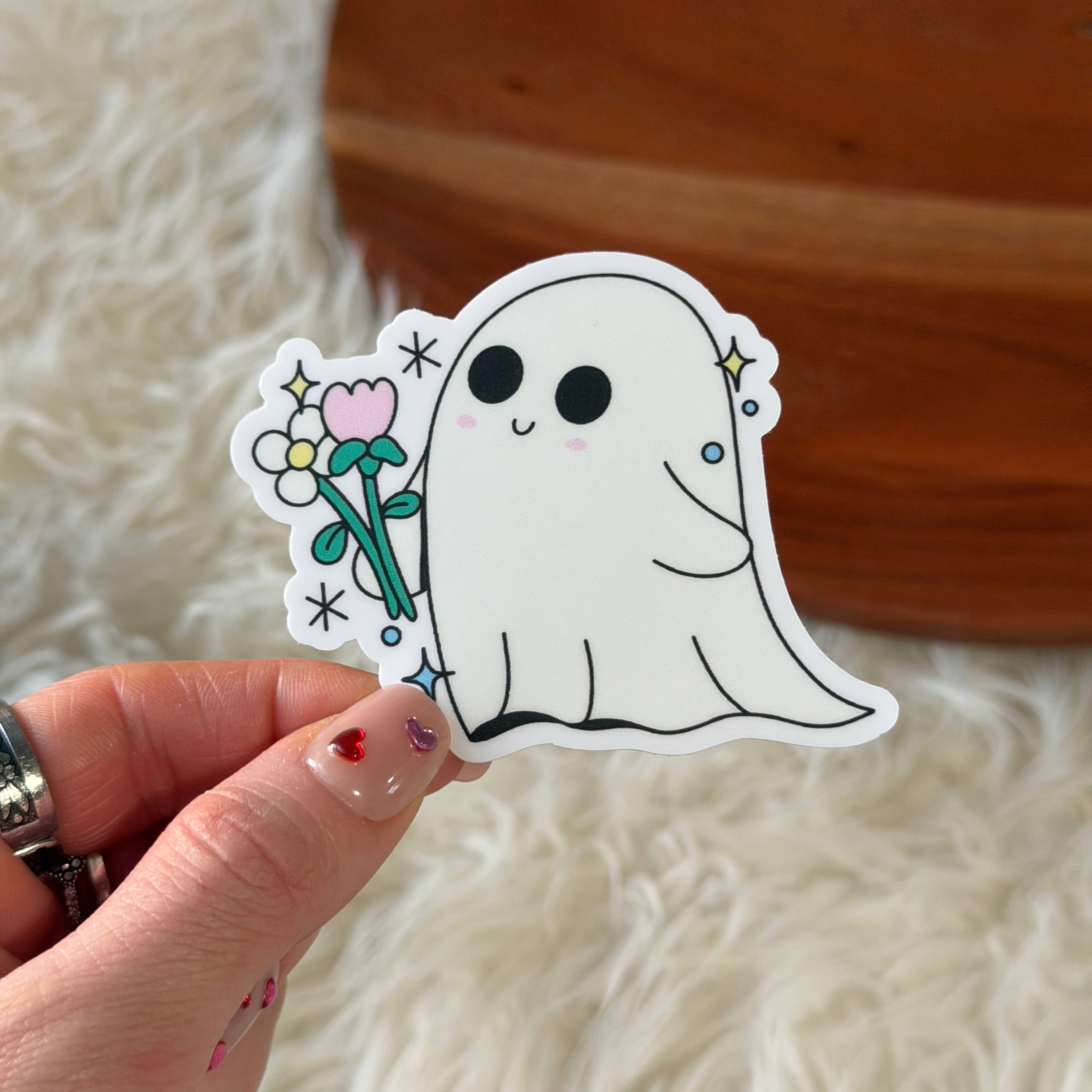 A hand holds a die-cut sticker featuring a cute white cartoon ghost holding a pink and yellow flower bouquet, set against a soft white faux fur background with a wooden surface behind it.