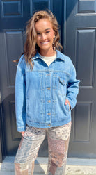 A woman wearing a light blue denim jacket over a white t-shirt and patterned pants, standing in front of a dark blue paneled door.