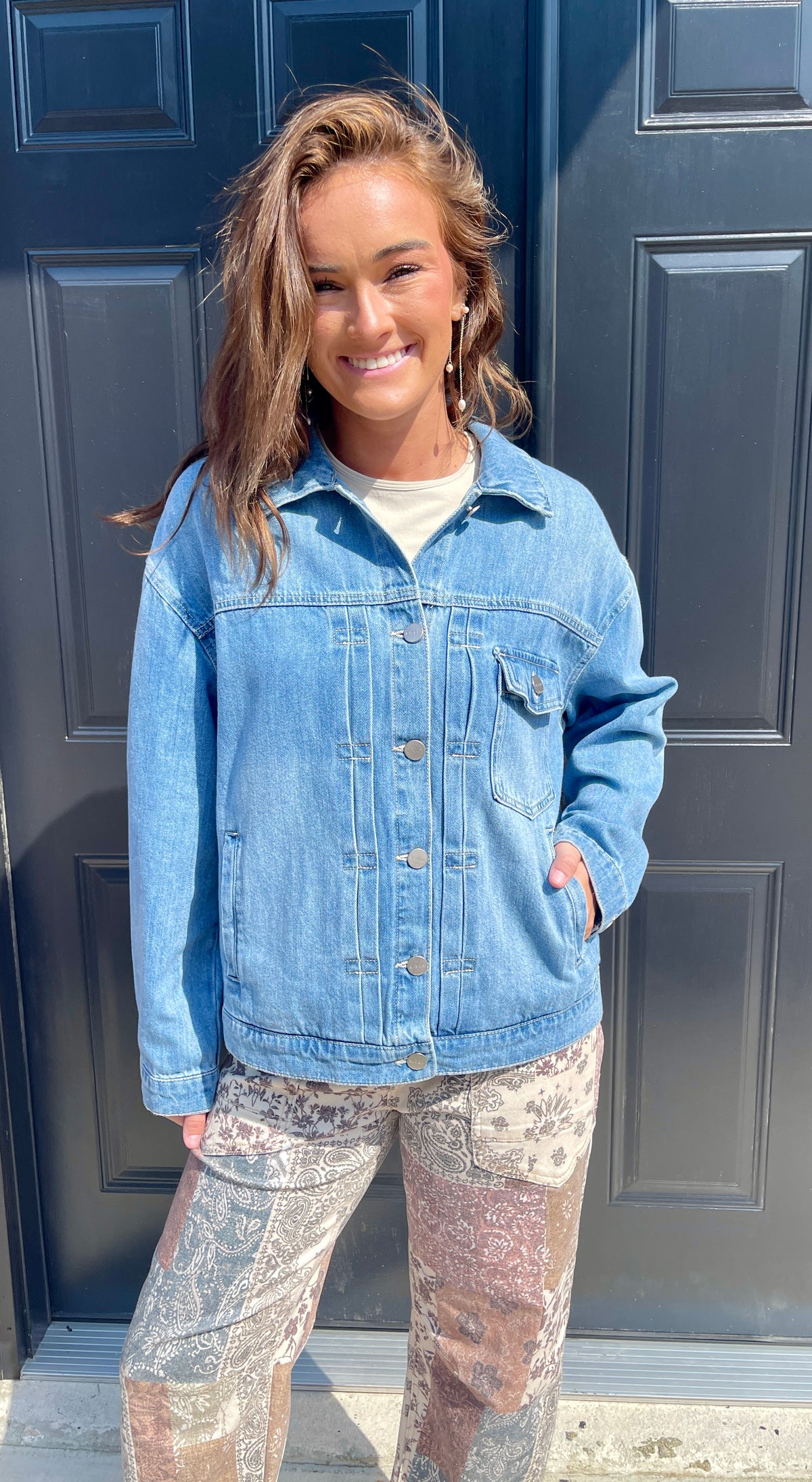 A woman wearing a light blue denim jacket over a white t-shirt and patterned pants, standing in front of a dark blue paneled door.