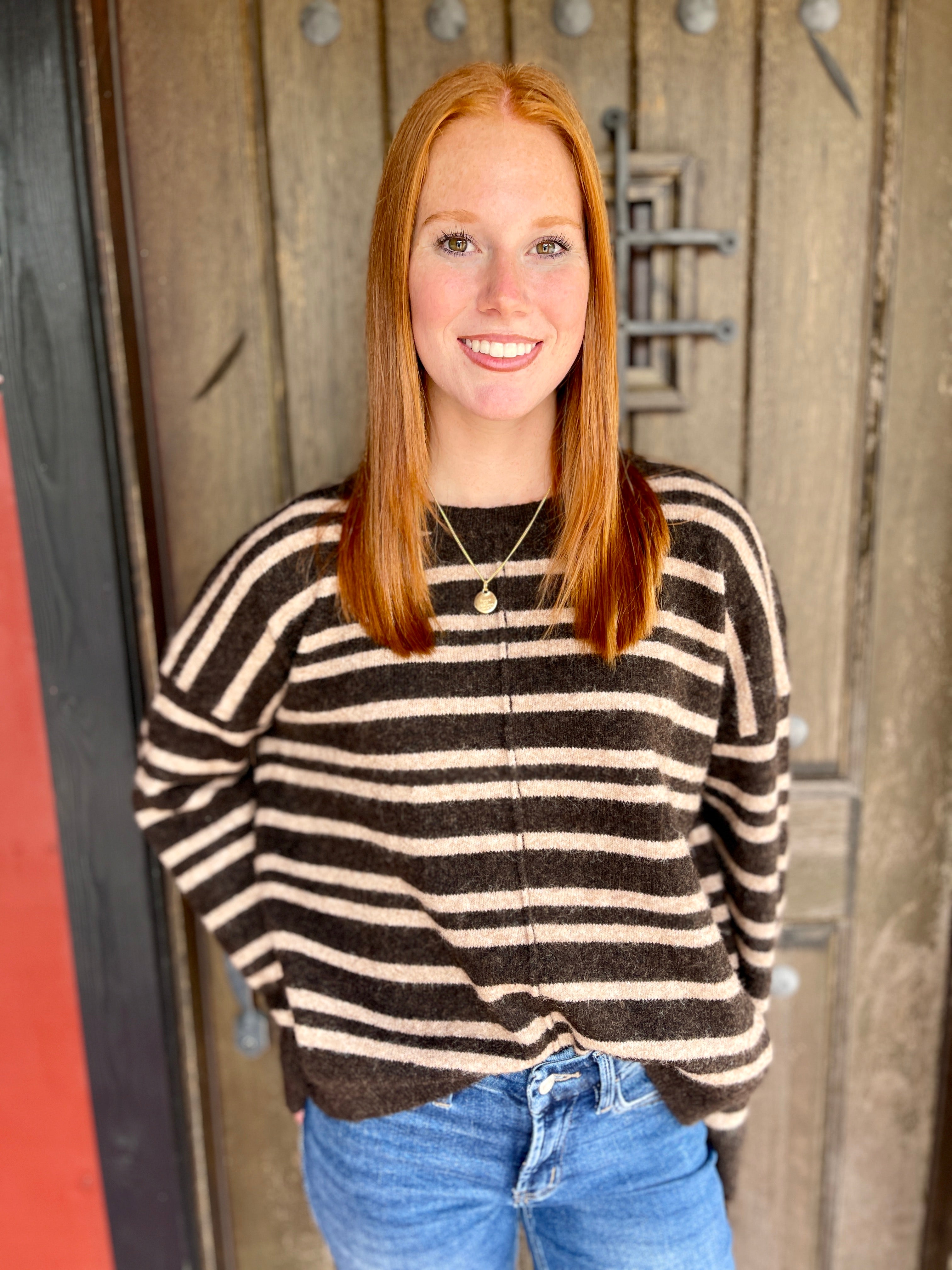 A smiling young woman with red hair stands in front of a rustic wooden door, wearing a brown and cream striped knit sweater and blue jeans.