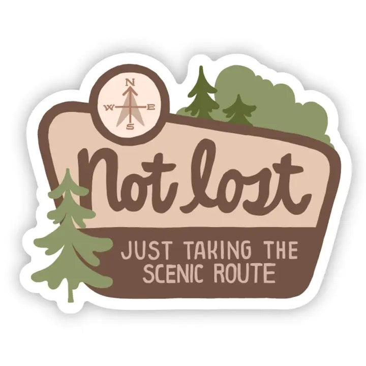 A die-cut sticker with a white border featuring a brown and beige sign design. The sign reads 'Not lost' with a compass rose at the top and 'JUST TAKING THE SCENIC ROUTE' below.