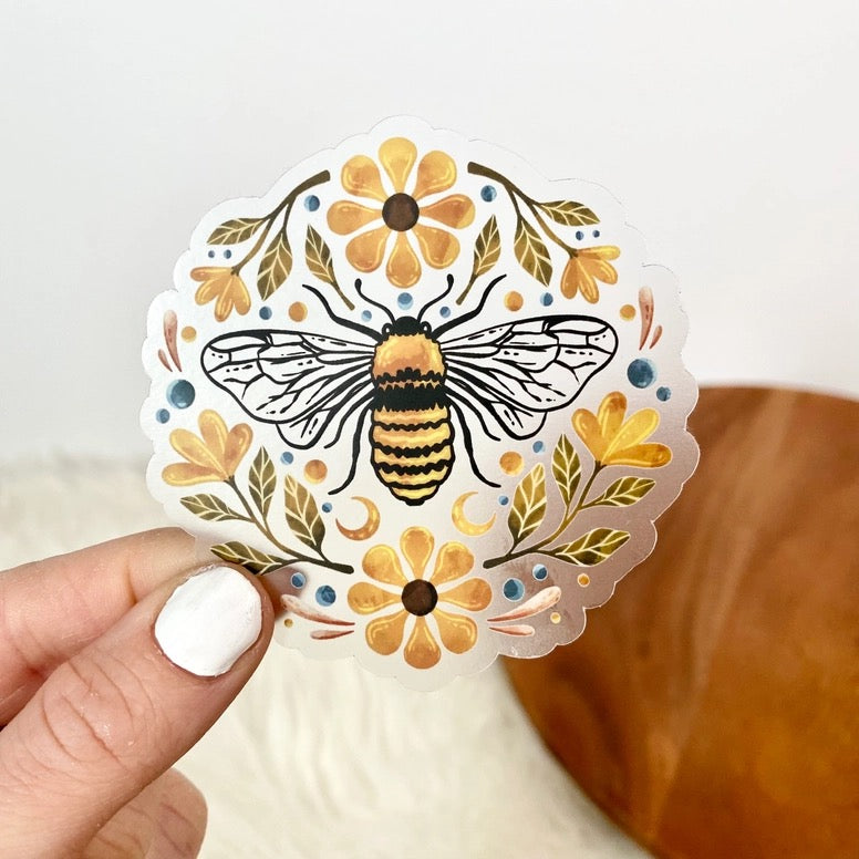 Decorative sticker with a bee and flowers held by a hand against a neutral background