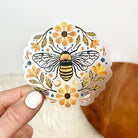Decorative sticker with a bee and flowers held by a hand against a neutral background