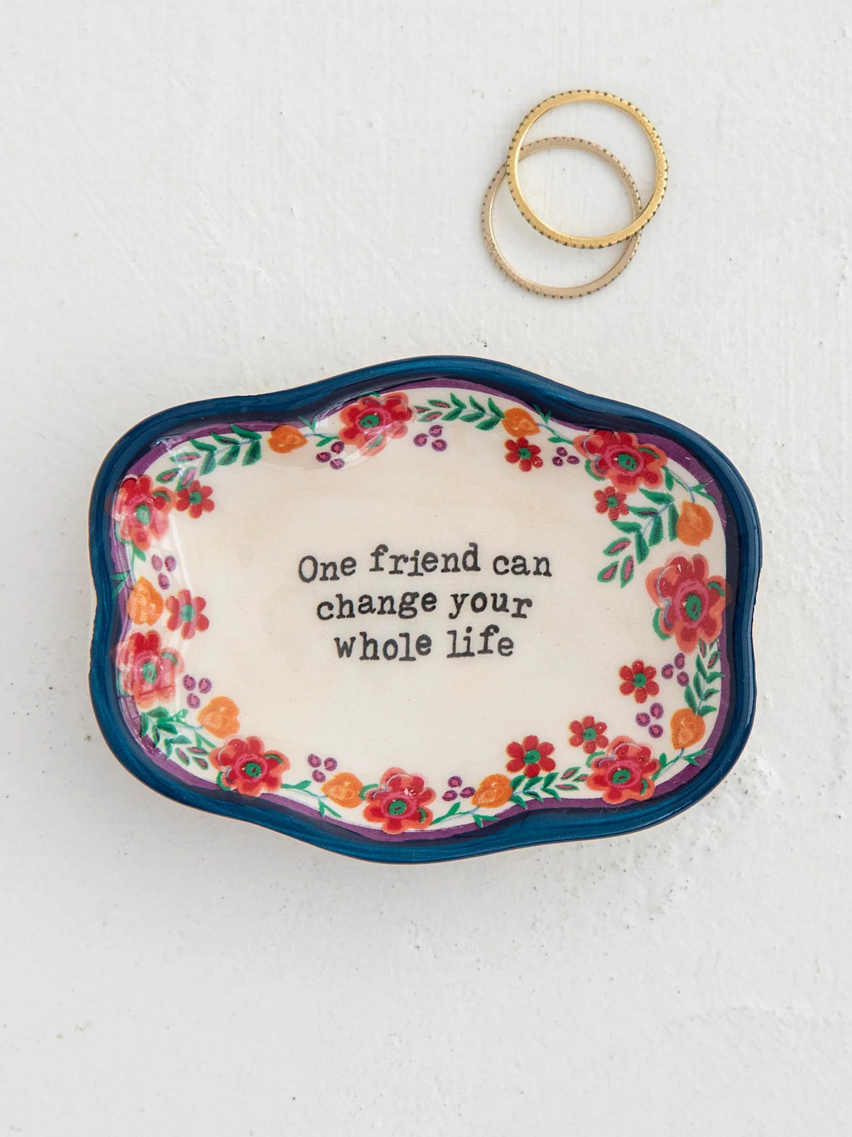 DECORATIVE BOWL WITH ONE FRIEND CAN CHANGE YOUR WHOLE LIFE WITH FLOWERS AND NAVY RIM 2 RINGS SET TO THE SIDE ON A WHITE BACKGROUND