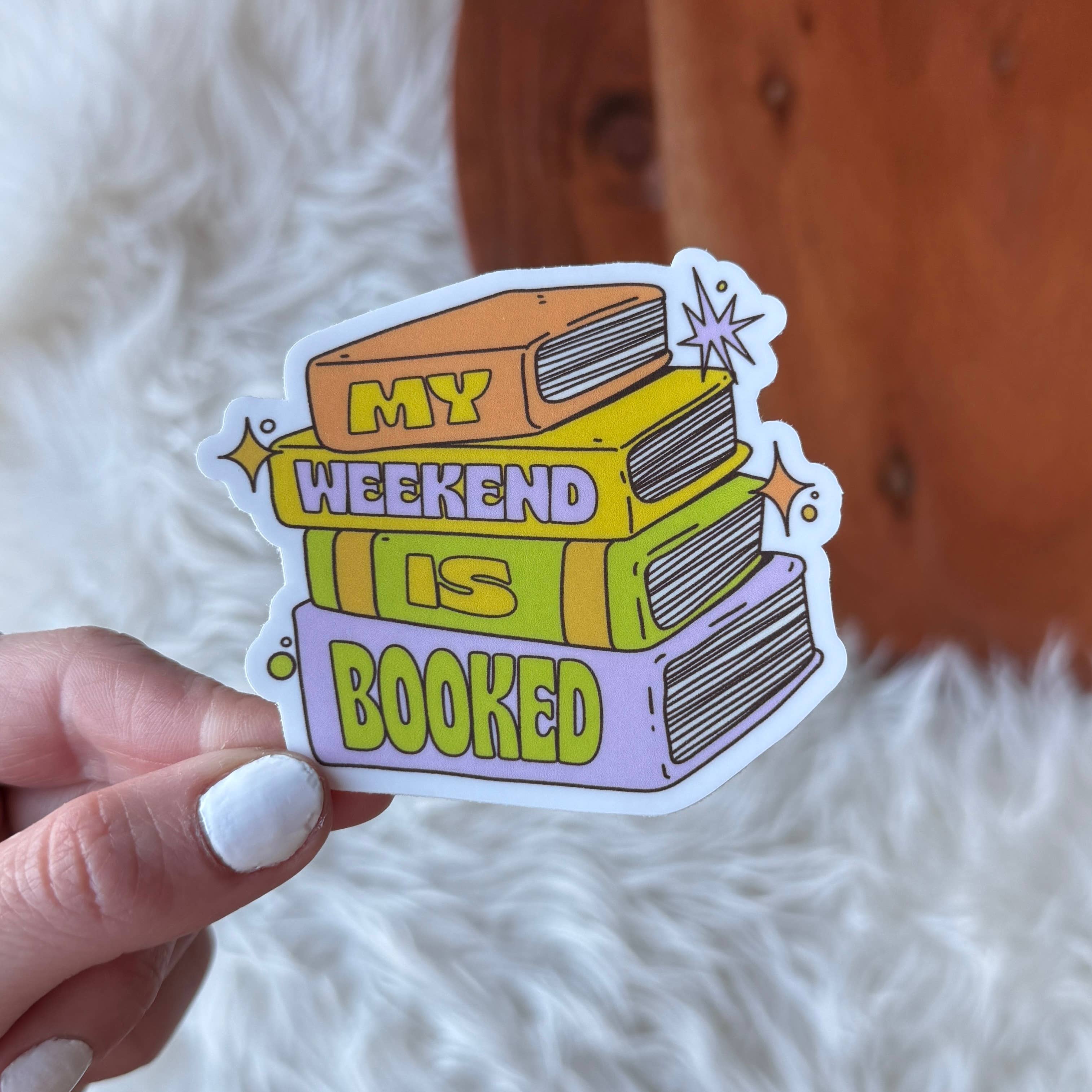 Sticker with books design and 'My weekend is booked' text held by a hand on a soft surface.