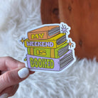 Sticker with books design and 'My weekend is booked' text held by a hand on a soft surface.