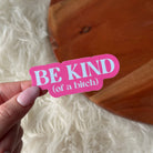 Pink sticker with 'BE KIND (of a bitch)' text held by a hand against a wooden surface.