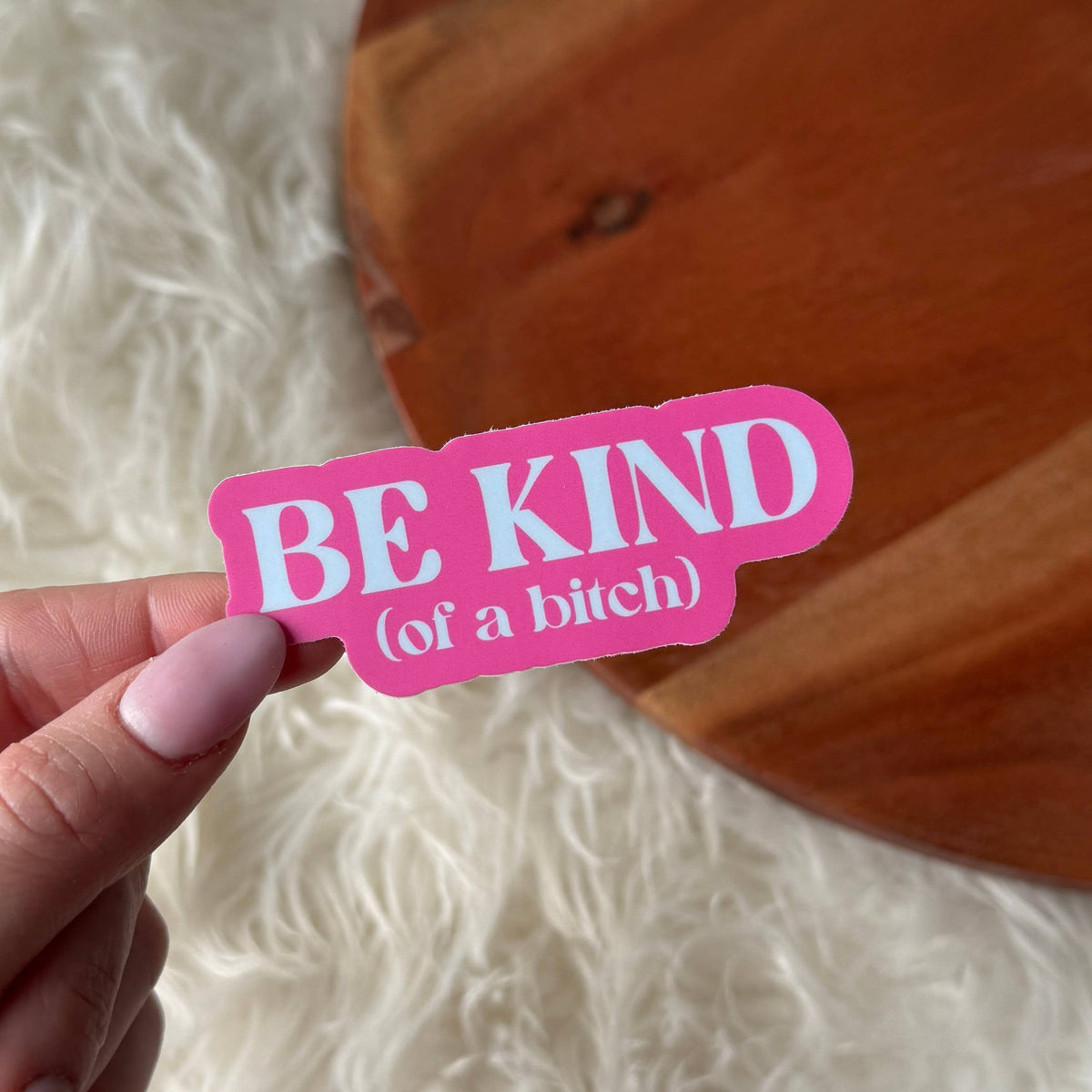 Pink sticker with 'BE KIND (of a bitch)' text held by a hand against a wooden surface.